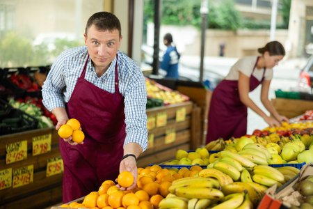 Male seller of vegetable department of store is replenishing showcase with orangesの写真素材