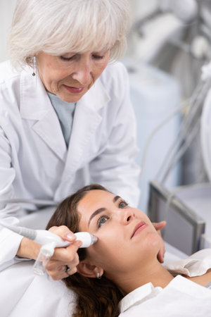 Young woman at appointment with beautician doing ultrasound face cleaning with help of high-frequency ultrasonic waves in cosmetology clinicの写真素材