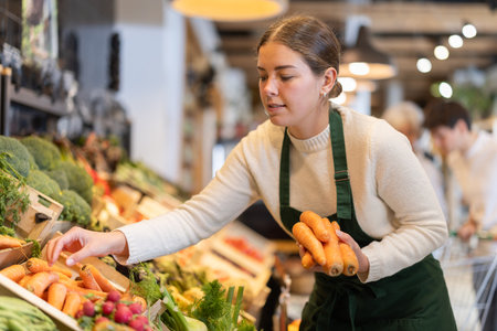 Girl salesperson adjusts carrot vegetables in stores display caseの写真素材