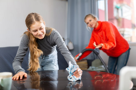 Hardworking daughter cleans the table with a rag while mother vacuums roomの写真素材