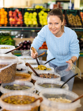Middle-aged woman purchaser putting olives into container in grocery storeの写真素材