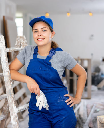 Portrait of contented young female builder in blue overalls during the repair of cottage