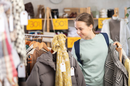 Positive young girl standing in interior of clothing boutique and choosing something on sale of winter clothing collectionの写真素材