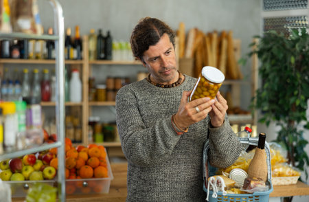 Man customer with shopping cart or basket buying food in grocery store, reading ingredients on jar of conserve olivesの写真素材