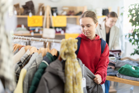 Positive young girl standing in interior of clothing boutique and choosing something on sale of winter clothing collectionの写真素材