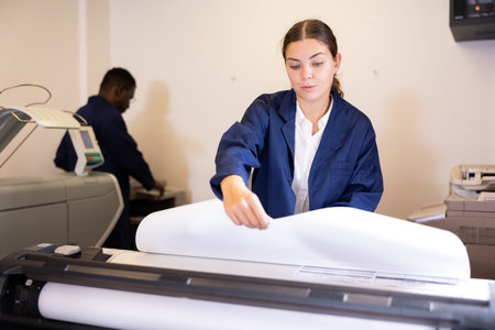 Concentrated young female in a blue robe uniform using plotter while printing a banner in the...