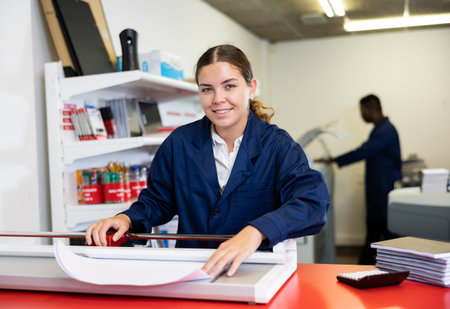 Joyful young woman in a blue uniform smiling at the camera and using paper cutter in printer house