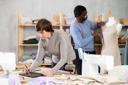 Woman tailor creates sketch of dress on tablet, man companion work in background with mannequin