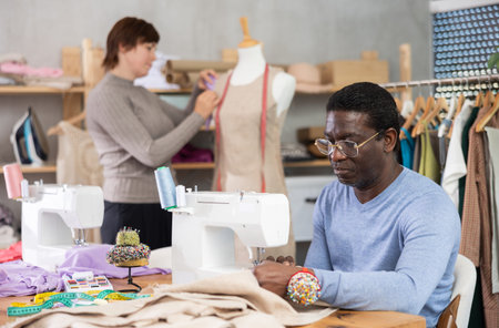 African American man tailor sews clothes, companion work in background with mannequin