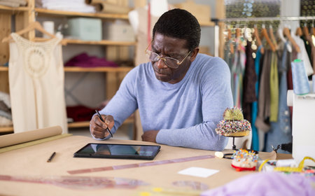 Male tailor draws sketch of future clothing on an electronic tablet. Working with modern gadgets in sewing workshopの写真素材