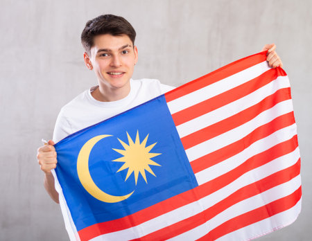 Man in casual clothes holds unfurled flag of Malaysia in hands raised above head against gray wallの写真素材