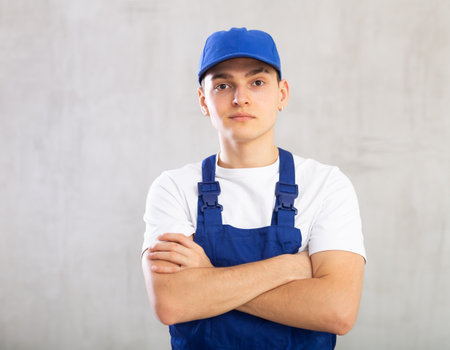 Positive male worker in blue overall and cap poses with folded arms on chest on gray backgroundの写真素材