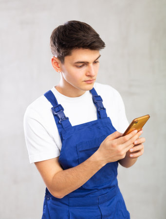 Young male builder in protective overalls with notebooks in his hands writes a message on smartphone screenの写真素材