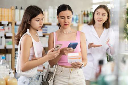 Portrait of mother and daughter choosing box with pills to improve well-being in pharmacy, scan QR code on label, young woman pharmacist consulting customersの写真素材