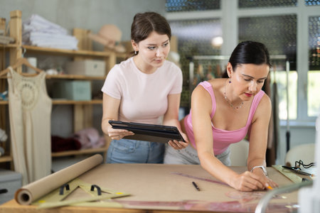 Two female dressmakers draw outline of dress on paper while looking at the screen of an electronic tabletの写真素材
