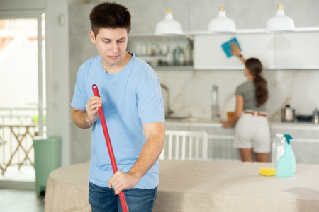 Guy mopping the floor in the kitchen while his wife is cleaning the kitchen-cupboard