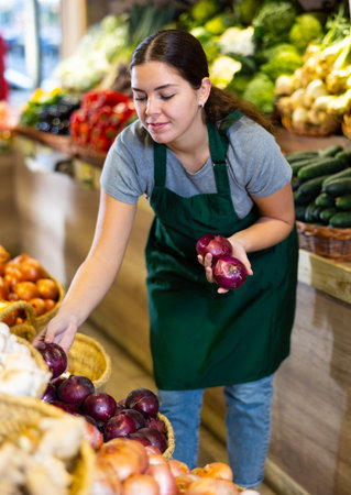 Female grocery store worker arranges red onion and other vegetables on counter and display caseの写真素材