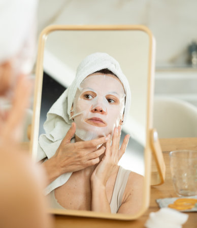 Woman applying moisturizing sheet face mask in front of mirrorの写真素材