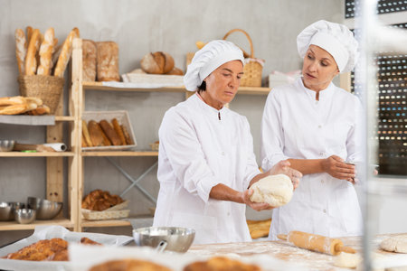 Elderly female baker giving advice to young assistant in kitchen