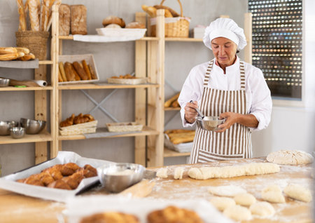 Portrait of happy mature female professional baker in uniform standing and posing in bakery during...