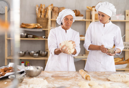 Two skilled female bakers in white uniform kneading and cutting dough, preparing portioned pieces before baking in bakehouseの写真素材