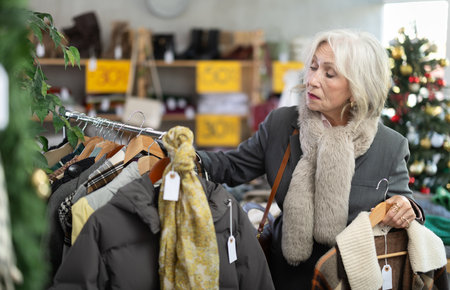 Attentive mature woman choosing coat or jacket in clothing storeの写真素材