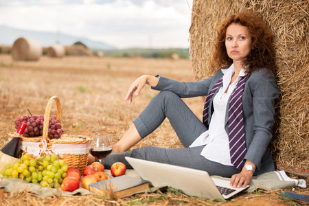 Portrait of woman in office clothes with laptop on picnic at farm fieldの写真素材