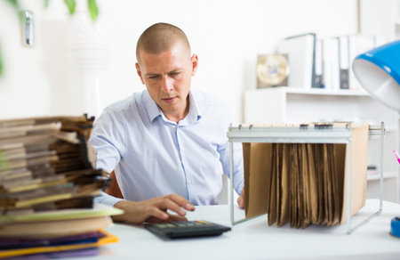 Accountant counts on calculator and works with documents in file cabinet modern officeの写真素材