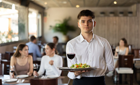 Male waiter in uniform posing with tray and glass of red wine in his hands in restaurant