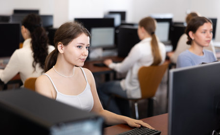 Young woman student of computer courses looks at monitor screen, types on keyboard and does work