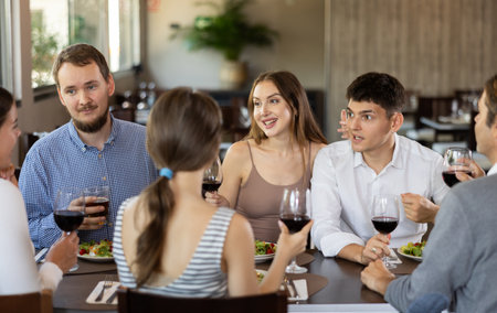 Group of positive friends, men and women, are sitting at a table in a restaurant. They are chatting and drinking wineの写真素材
