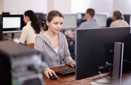 Young woman student of computer courses looks at monitor screen, types on keyboard and does workの写真素材