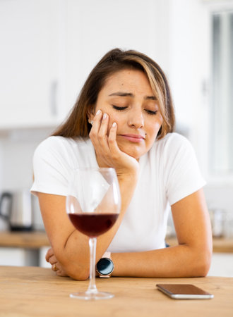 Frustrated young woman drinking wine and waiting for call in home kitchenの写真素材