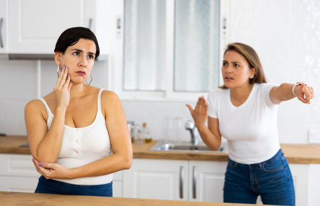 Upset young woman standing in home kitchen while sister reprimanding herの写真素材