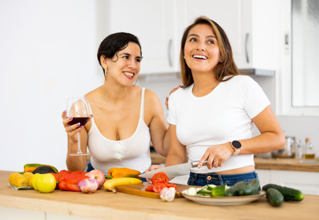 Happy female friends with glasses of red wine chatting and preparing salad together in kitchenの写真素材