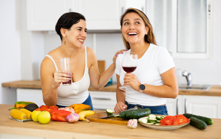 Two cheerful female roommates drinking wine and preparing dinner at homeの写真素材