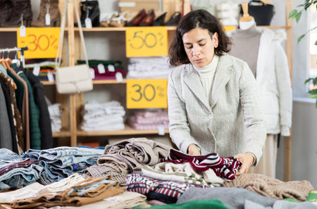 European female buyer standing in clothing market and carefully examines selection of warm sweaters for winter seasonの写真素材