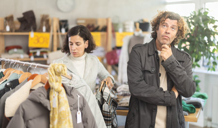European female customer chooses new jacket or coat for winter season, man bored while waiting for...