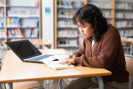 Portrait of focused teenage female pupil sitting at desk studying in library with classmatesの写真素材