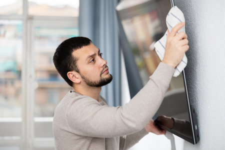 Young man rubbing screen of wall-mounted TV in apartmentの写真素材