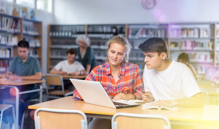 Concentrated teenager female schoolgirl and her male friend sitting at library desk, doing homework...