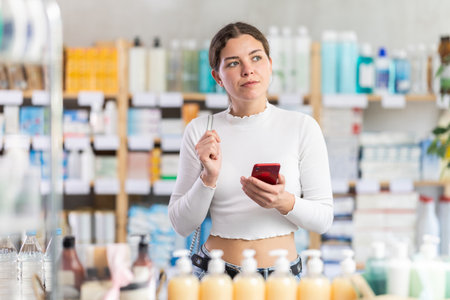 Female shopper searches for necessary goods on the shelves of a supermarket, checking the list on smartphoneの写真素材