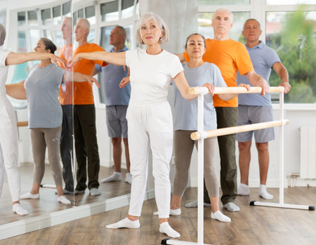 Dancing positive elderly women and men engaged in group class perform exercise near ballet barre, standing in 2nd position of ballet stance in dance studioの写真素材