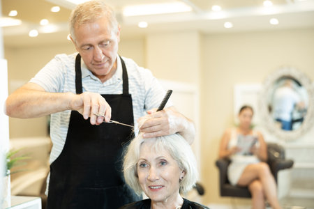 Elderly hairdresser using scissors while providing haircut to woman in salonの写真素材