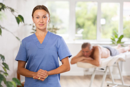 Positive middle-aged masseuse posing against background of therapy roomの写真素材