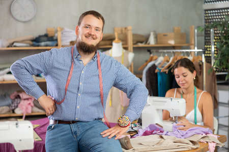 Male dressmaker welcoming clients while young seamstress is sewing behindの写真素材