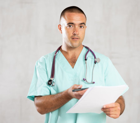 Portrait of male doctor in medical gown with stethoscope making notes in patient medical historyの写真素材