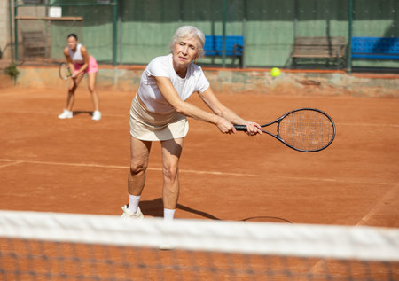 Sporty two women during doubles match on tennis court. Active old lady hitting tennis ball with racketの写真素材