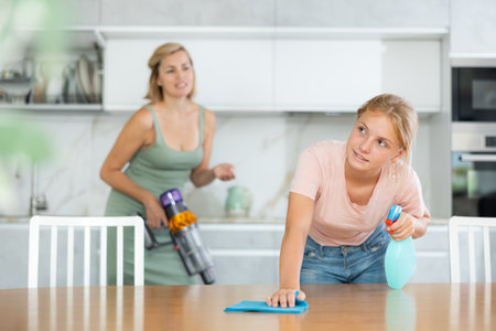 Teen girl help mother and wash dining table in kitchen, blurry mom cleaning using vacuum cleanerの写真素材