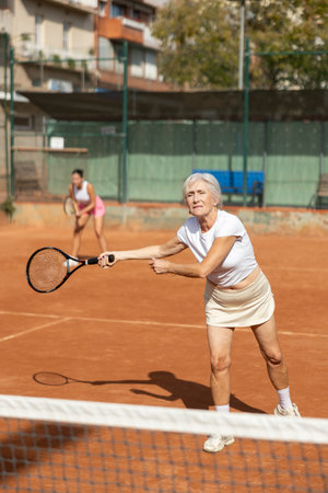 Sporty two women during doubles match on tennis court. Active old lady hitting tennis ball with racketの写真素材